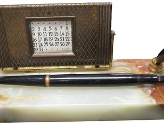 Op de afbeelding: Een vintage bureauset van marmer met een vergulde kalender en een zwarte vulpen. De kalender heeft een klein venster dat de datum en de dagen van de week weergeeft. De pen heeft een zwarte huls en een vergulde punt.