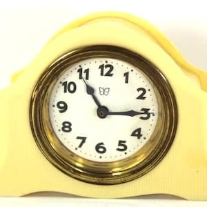 May include: A cream-colored, vintage-style clock with a gold-tone bezel and a white face. The clock has black numerals and hands, and a small butterfly emblem. The time displayed is approximately 2:55. The clock is made of a light yellow material.