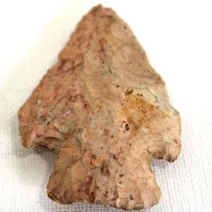 以下が含まれることがあります： 茶色とベージュの矢じりで、三角形の頭と切り込みの入った基部があります。 石の遺物はざらざらした質感で、白い背景に置かれています。