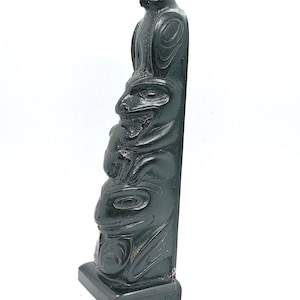 Könnte beinhalten: Dunkelgrüne Totempfahl-Skulptur mit geschnitzten Tierfiguren, darunter ein Vogel an der Spitze. Der Totempfahl ist rechteckig mit quadratischer Basis, vor weißem Hintergrund.