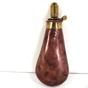 Puede incluir: Antigua petaca de pólvora de cobre con detalles de latón. La petaca tiene forma de lágrima y una parte superior de latón con una palanca. El cobre presenta una rica pátina envejecida.