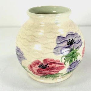 May include: A small, round ceramic vase with a cream-colored base and a green rim. The vase is decorated with hand-painted flowers in shades of red and purple, with green leaves. The vase has a wavy pattern.