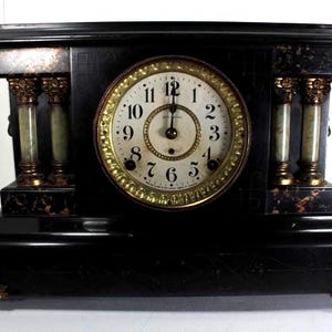 May include: An antique black mantel clock with a round clock face. The clock has a gold bezel and Roman numerals. Decorative columns with green and gold accents are visible. The base has gold-coloured feet.