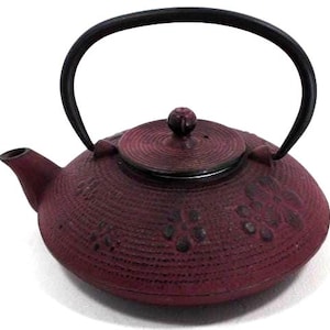 Peut inclure: Une théière en fonte de couleur bordeaux avec une anse et un couvercle noirs. La théière a une surface texturée avec des motifs floraux. La poignée est arquée sur le dessus et le bec est sur le côté gauche.
