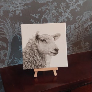 Può includere: Un disegno in bianco e nero di una testa di pecora su una piastrella bianca, esposto su un piccolo cavalletto di legno.