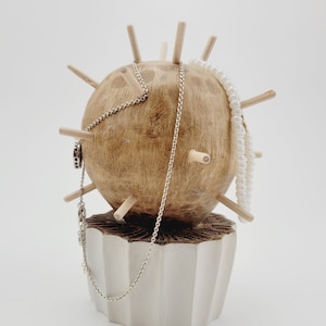 Può includere: Un porta gioielli in legno a forma di sfera con più punte di legno che sporgono dalla superficie. Il porta gioielli ha una base in ceramica bianca e tiene una collana a catena d'argento e una collana di perle bianche.