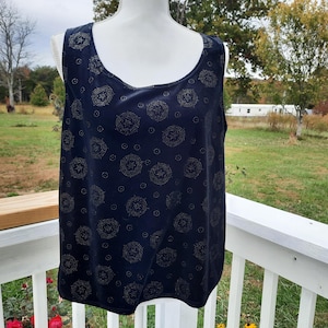 May include: A navy blue tank top with a scoop neckline. The top is decorated with a repeating pattern of gold-colored circular designs. The sleeveless top is displayed on a mannequin against a backdrop of a green lawn and trees.