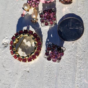 May include: A collection of vintage jewelry pieces, including a gold-toned brooch with red gemstones, a pair of square earrings, and a cluster of earrings with a coin for size comparison. The jewelry features pink and red stones.