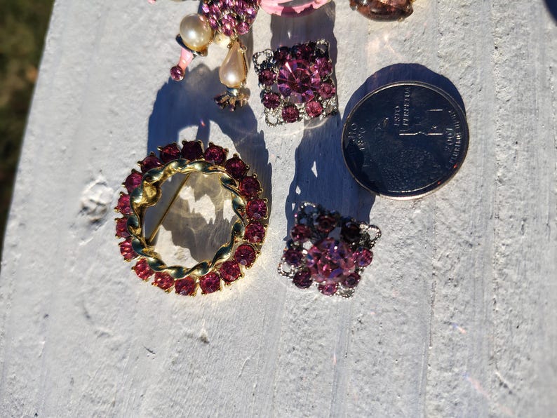 May include: A collection of vintage jewelry pieces, including a gold-toned brooch with red gemstones, a pair of square earrings, and a cluster of earrings with a coin for size comparison. The jewelry features pink and red stones.