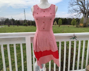 Könnte beinhalten: Ein ärmelloses rot-weißes Gingham-Kleid mit rundem Ausschnitt und eng anliegendem Oberteil. Das Kleid hat kleine rote Blumenakzente und einen roten Einsatz am Saum. Das Kleid wird auf einer Schaufensterpuppe präsentiert.