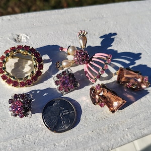 May include: A collection of vintage jewelry pieces, including a circular brooch with red stones, a butterfly brooch with pink wings, square and rectangular earrings, and a coin. The jewelry is set on a white surface.
