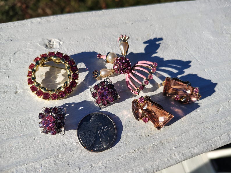 May include: A collection of vintage jewelry pieces, including a circular brooch with red stones, a butterfly brooch with pink wings, square and rectangular earrings, and a coin. The jewelry is set on a white surface.