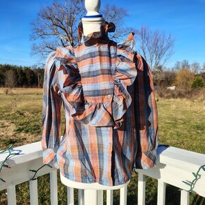 May include: A vintage-style blouse with a plaid pattern in shades of orange, blue, and black. The blouse features long sleeves, a ruffled collar, and a button-down front. The garment is displayed on a mannequin outdoors with a natural background.