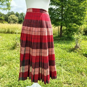 May include: A high-waisted, pleated midi skirt featuring a bold plaid pattern in shades of deep red, muted pink, and charcoal gray. The skirt has a structured waistband and a full, flowing silhouette that falls below the knee with vertical pleats.