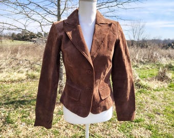 May include: A brown suede blazer with a notched lapel, two front pockets, and a two-button closure. The jacket is displayed on a white mannequin against a natural outdoor backdrop.