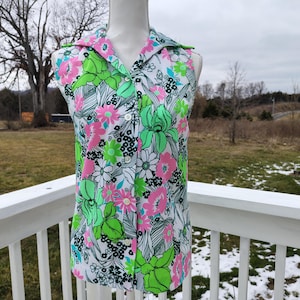 May include: A sleeveless, button-down top with a floral pattern. The white fabric is covered in large pink and green flowers, with black accents. The top has a collar and is displayed on a mannequin.