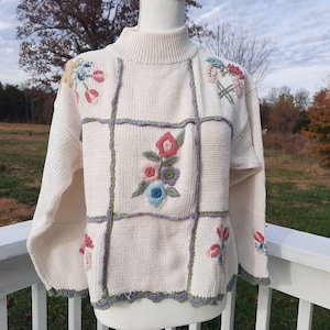 May include: Cream-colored sweater with a turtleneck and floral embroidery. The sweater features a patchwork design with embroidered flowers in pink, blue, and purple. The edges are trimmed with a green and gray scalloped design.