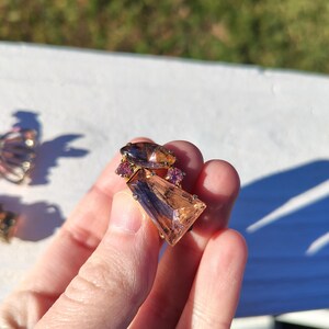 May include: A vintage brooch featuring a large, rectangular, peach-colored gemstone with a dark brown accent. The brooch is accented with smaller, round, pink gemstones and a dark brown, oval-shaped stone. The setting appears to be gold-toned.