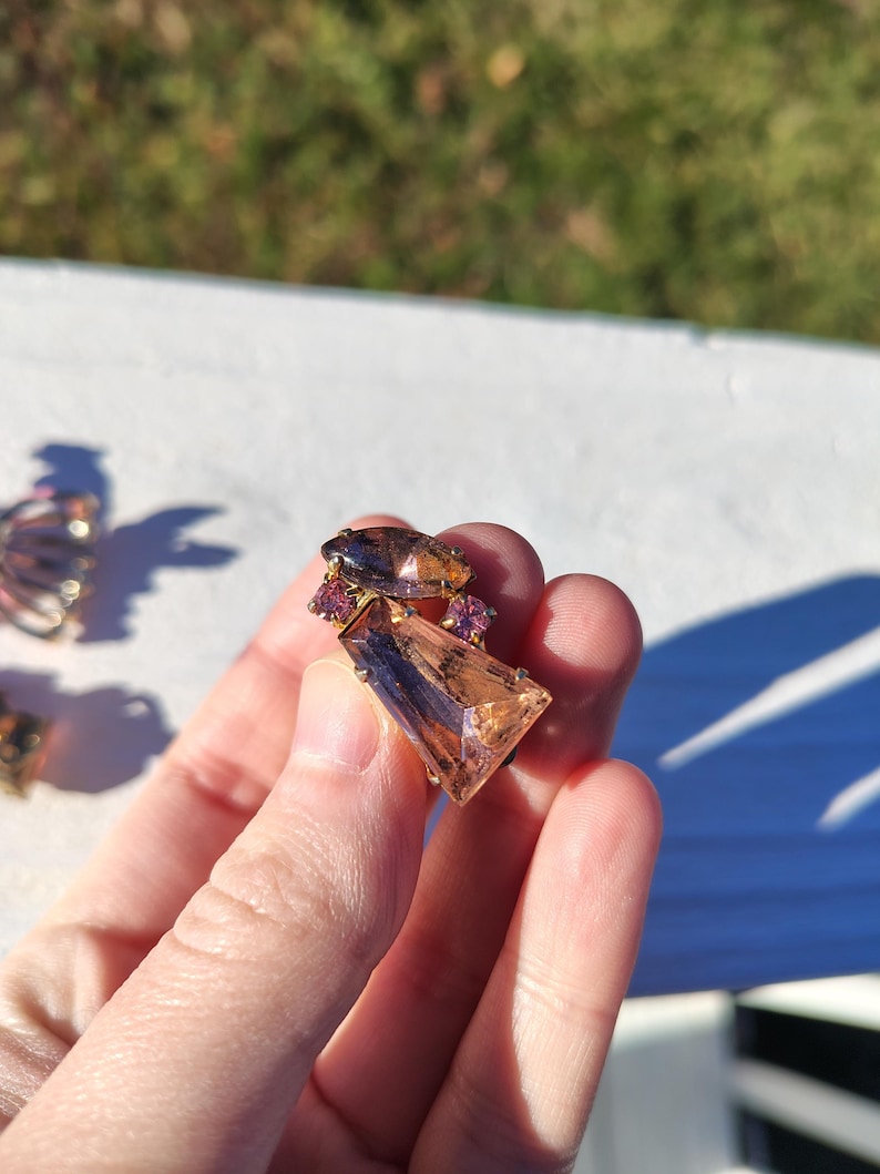 May include: A vintage brooch featuring a large, rectangular, peach-colored gemstone with a dark brown accent. The brooch is accented with smaller, round, pink gemstones and a dark brown, oval-shaped stone. The setting appears to be gold-toned.