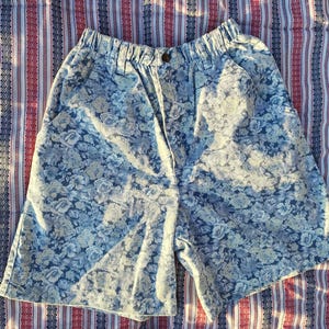 May include: Light blue floral print shorts with an elastic waistband and button closure. The shorts feature a faded floral pattern in shades of blue and white. The shorts are made of a lightweight fabric.