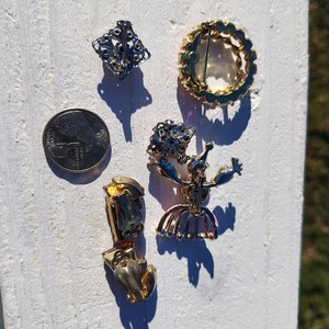May include: An assortment of vintage jewelry pieces, including a silver-toned square brooch, a gold-toned circular brooch, a gold-toned ballerina brooch, and a pair of gold-toned clip-on earrings. A U.S. quarter is included for size reference. The jewelry is displayed on a white surface.