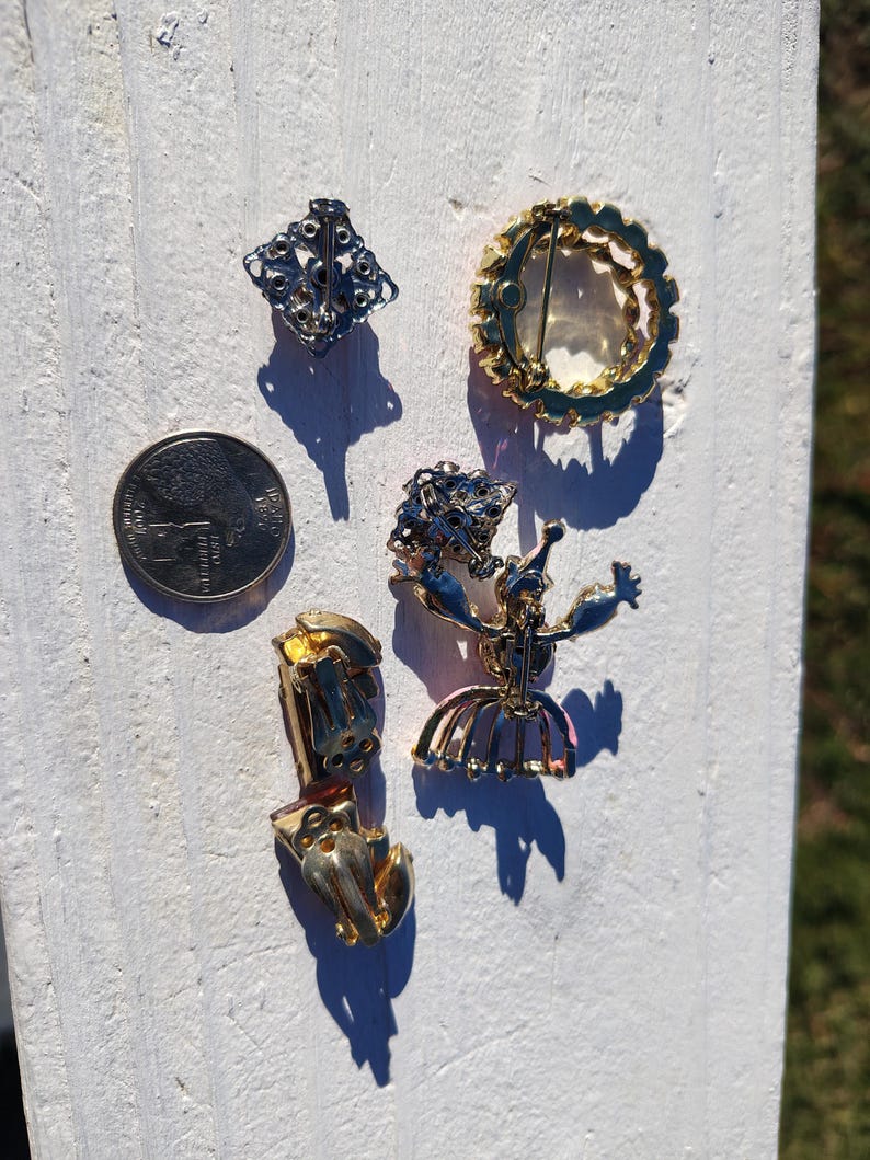 May include: An assortment of vintage jewelry pieces, including a silver-toned square brooch, a gold-toned circular brooch, a gold-toned ballerina brooch, and a pair of gold-toned clip-on earrings. A U.S. quarter is included for size reference. The jewelry is displayed on a white surface.