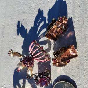 May include: An assortment of vintage jewelry pieces, including clip-on earrings and a brooch. The earrings feature rectangular, amber-colored stones and small, round, purple gems. The brooch has a pink and black design with purple gems and faux pearls.