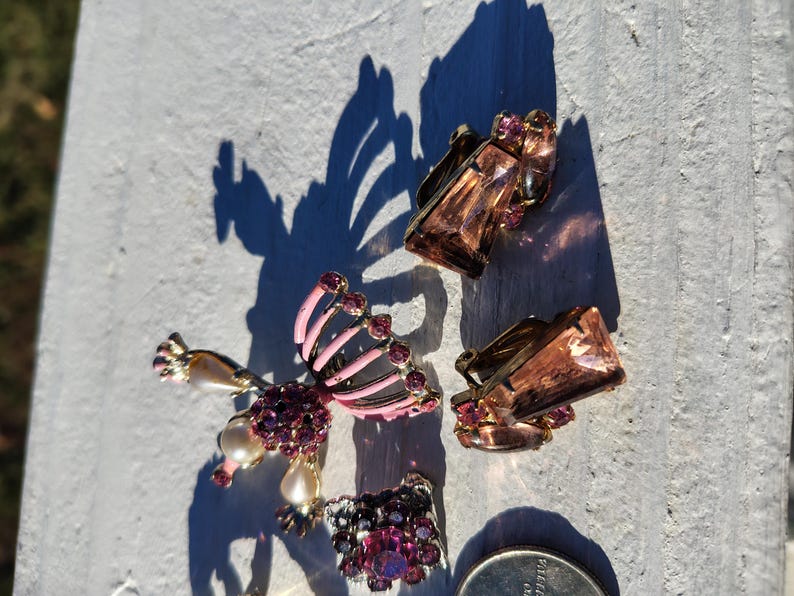 May include: An assortment of vintage jewelry pieces, including clip-on earrings and a brooch. The earrings feature rectangular, amber-colored stones and small, round, purple gems. The brooch has a pink and black design with purple gems and faux pearls.