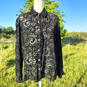 May include: Black button-down shirt with a detailed paisley pattern. The long-sleeved shirt has a classic collar and is displayed on a mannequin. The shirt is made of a sheer fabric, allowing the mannequin to be seen through the shirt.