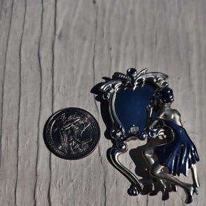 May include: A silver-toned metal brooch depicting a figure in a dress looking into an ornate, oval-shaped mirror. The brooch is positioned next to a United States quarter coin for scale, which has a diameter of approximately 0.955 inches.
