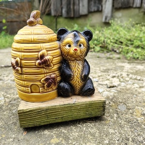 May include: A ceramic honey pot shaped like a beehive with bee accents, next to a bear figurine. The honey pot is yellow with brown details, and the bear is black and yellow. The items are on a green wooden block.