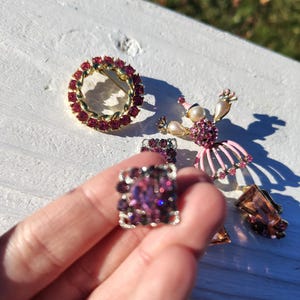 May include: A collection of vintage jewelry pieces, including a round brooch with red stones, a square ring with purple stones, and a ballerina brooch with pink accents. Other pieces include earrings with brown stones.