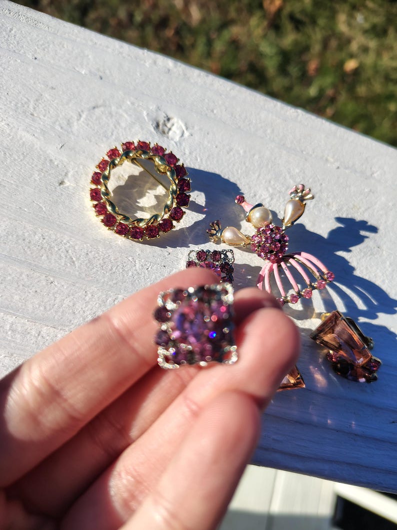 May include: A collection of vintage jewelry pieces, including a round brooch with red stones, a square ring with purple stones, and a ballerina brooch with pink accents. Other pieces include earrings with brown stones.