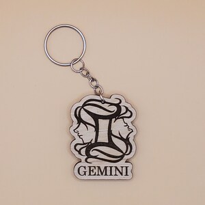 May include: A wooden keychain with a black and white illustration of the Gemini zodiac sign. The keychain has a silver ring and the word "GEMINI" is printed below the illustration.