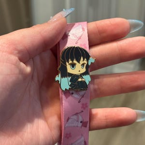 Puede incluir: Un cordón rosa con un patrón repetido de ilustraciones blancas y rosas y un pin dorado. El pin presenta un personaje de dibujos animados con cabello negro, detalles en azul y un atuendo negro. El cordón está sujeto por una mano.
