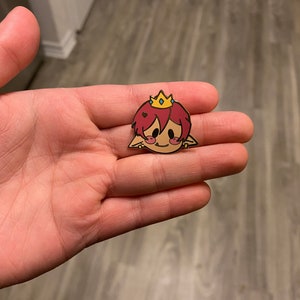 May include: A small, cartoon-style enamel pin featuring a character with red hair, a gold crown, and a smiling face. The pin is held in a hand, showcasing its size and design. The pin has a black outline.