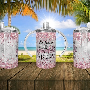 Op de afbeelding: Drie doorzichtige plastic bekers met handvatten en deksels. De bekers zijn gevuld met roze glitter. De middelste beker heeft de tekst "She leaves a little sparkle wherever she goes" erop gedrukt.