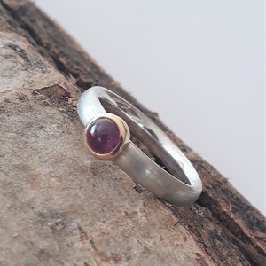 Könnte beinhalten: Ein Silberring mit einer goldenen Lünette, die einen runden, tiefroten Edelstein hält. Das Ringband hat eine gebürstete Oberfläche. Der Ring wird auf einem Holzstück präsentiert.