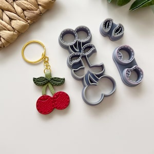 Puede incluir: Un juego de tres cortadores de galletas de plástico gris con forma de cerezas y hojas. Los cortadores se utilizan para crear galletas con forma de cereza u otros productos horneados. Los cortadores están sobre una superficie blanca.