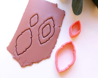 Può includere: Tre tagliabiscotti di plastica rosa a forma di foglie. I tagliabiscotti sono su una superficie bianca con un pezzo di argilla marrone dietro.