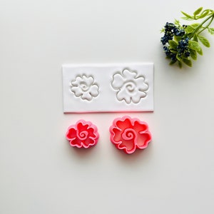 May include: Two pink plastic flower-shaped cookie cutters on a white surface. A white plastic sheet with two flower-shaped impressions is behind the cutters.