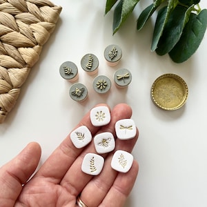 Könnte beinhalten: Satz von neun weißen Keramikstempeln mit Blattgold-Designs. Die Designs umfassen eine Biene, eine Libelle, eine Blume und Blätter. Die Stempel sind auf einer Hand und einer weißen Oberfläche angeordnet.