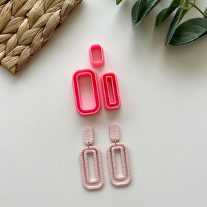 Op de afbeelding: Een set van drie roze plastic rechthoekige koekjesvormen en een paar roze dubbelgelaagde rechthoekige oorbellen met gouden accenten.