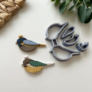 May include: Two felt bird shapes, one blue, brown, and yellow, the other green, yellow, and brown, with stitched details. A grey plastic cookie cutter in the shape of a bird.