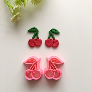 May include: Two sets of cherry cookie cutters. One set is red and green, the other is pink. Both sets are made of plastic and feature a cherry with a stem and leaf.