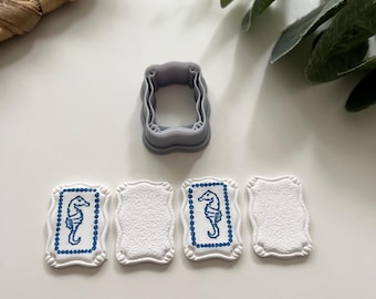 Pode incluir: Um cortador de biscoitos retangular cinza com uma borda ondulada está acima de quatro biscoitos brancos. Dois biscoitos apresentam um desenho de cavalo-marinho azul dentro de uma moldura pontilhada azul. Os outros dois são lisos.