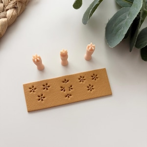 May include: A light brown leather stamp with a floral pattern and three small, light pink stamps. The stamps are arranged on a white surface with a woven texture and greenery in the background. The floral pattern is repeated on the leather stamp.