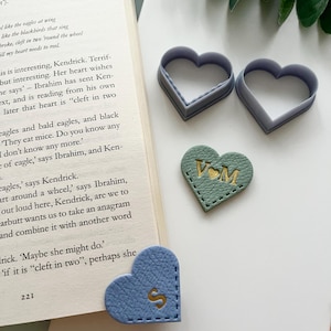 Op de afbeelding: Een assortiment hartvormige items. Twee grijze plastic hartvormige koekjessnijders, een groen leren hart met gouden letters en een blauwe leren hartvormige bladwijzer met een gouden letter "S".