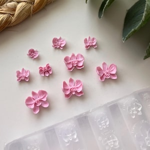 Puede incluir: Una colección de pequeños adornos rosas con forma de orquídea esparcidos sobre una superficie blanca. Se ve un molde transparente con formas de orquídeas. Las flores varían en tamaño y son de un rosa pastel suave.