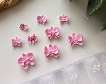 May include: A collection of small, pink orchid-shaped embellishments scattered on a white surface. A clear mold with orchid shapes is visible. The flowers vary in size and are a soft, pastel pink.